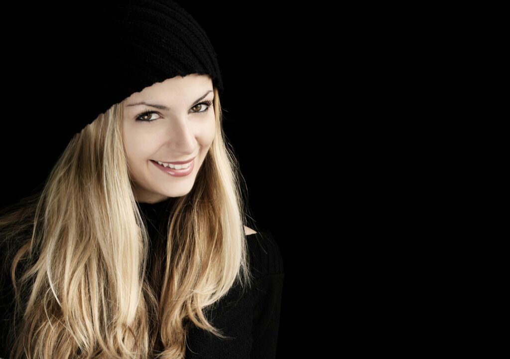 Beautiful woman with long blonde hair smiling in a black beanie against a dark background.
