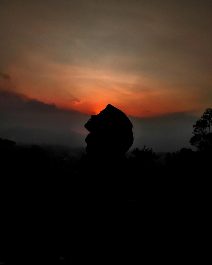 A serene silhouette of a person wearing a hijab against a vibrant sunset sky in a rural area.